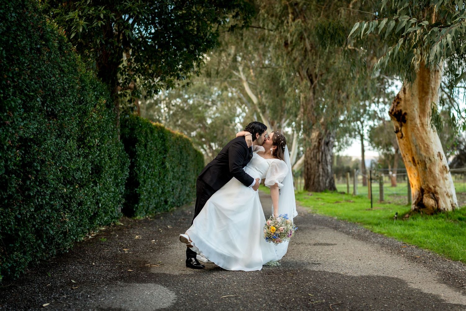 Bride and groom sharing a romantic dip kiss on a tree-lined garden path on their wedding day.