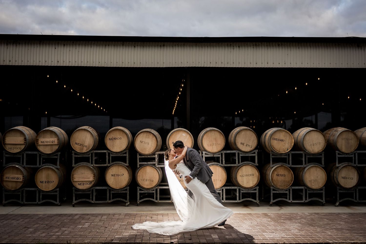 Couple shares romantic dip kiss in front of rustic wooden barrels at a winery wedding venue.