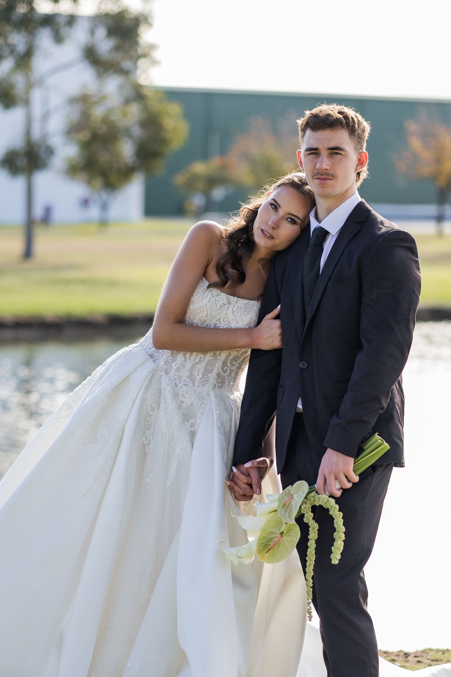 Young bride in white lace gown leans on groom in black suit holding bouquet by a pond.