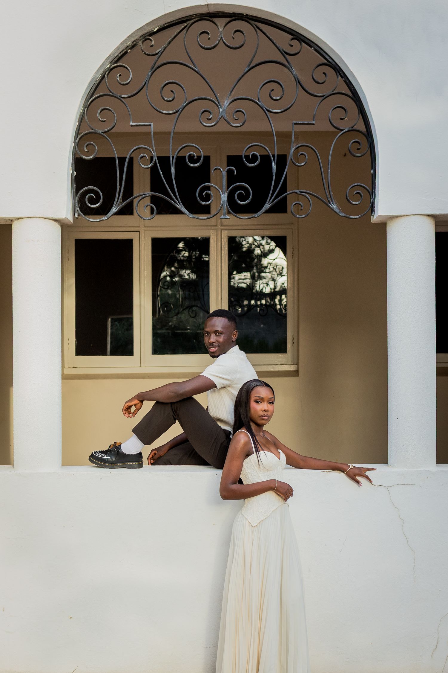 Couple posing on a white balcony ledge beneath an ornate arched window, he in a white shirt and she in a flowing cream gown.