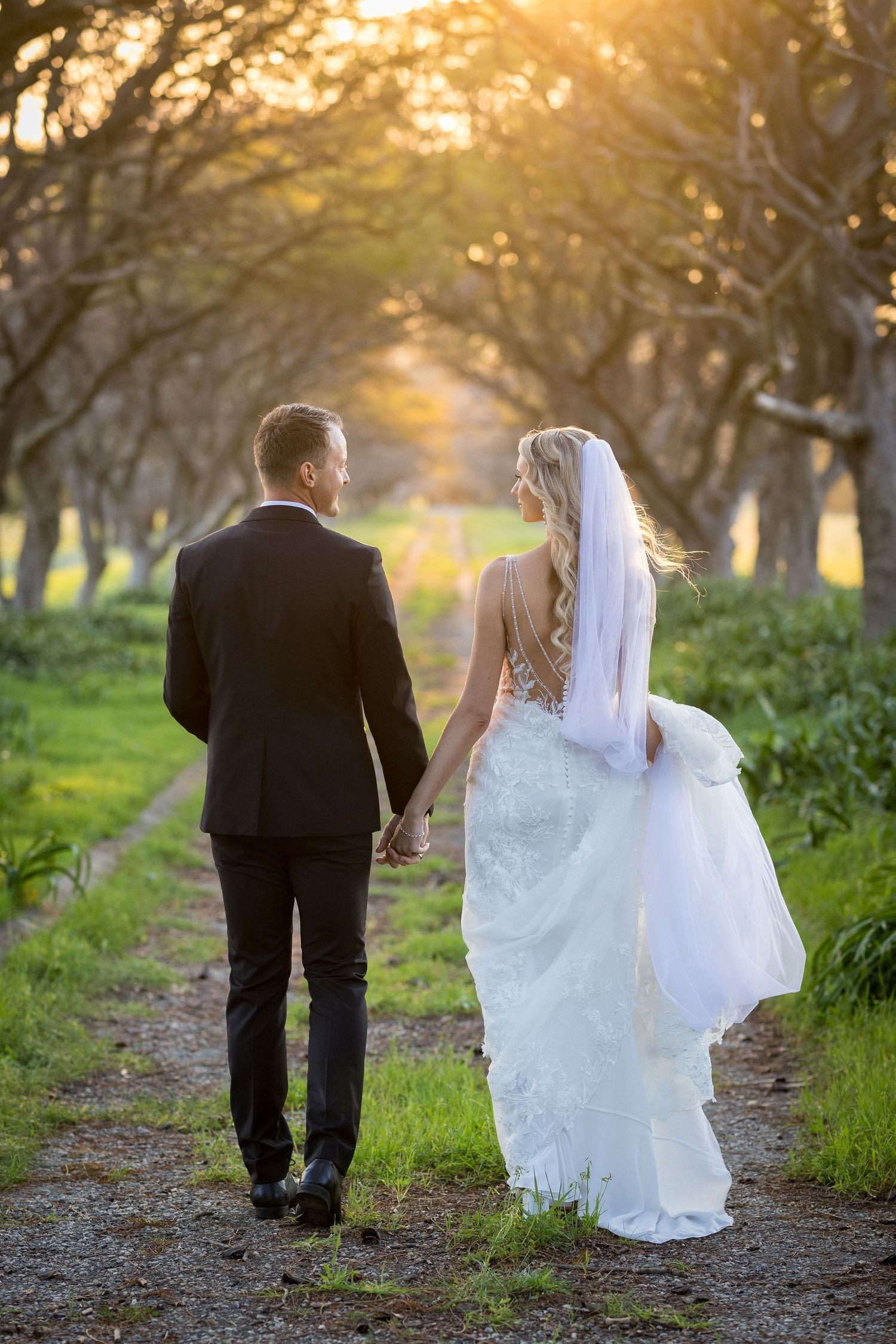 Bride and groom walk hand in hand down a tree-lined path bathed in golden sunset light.