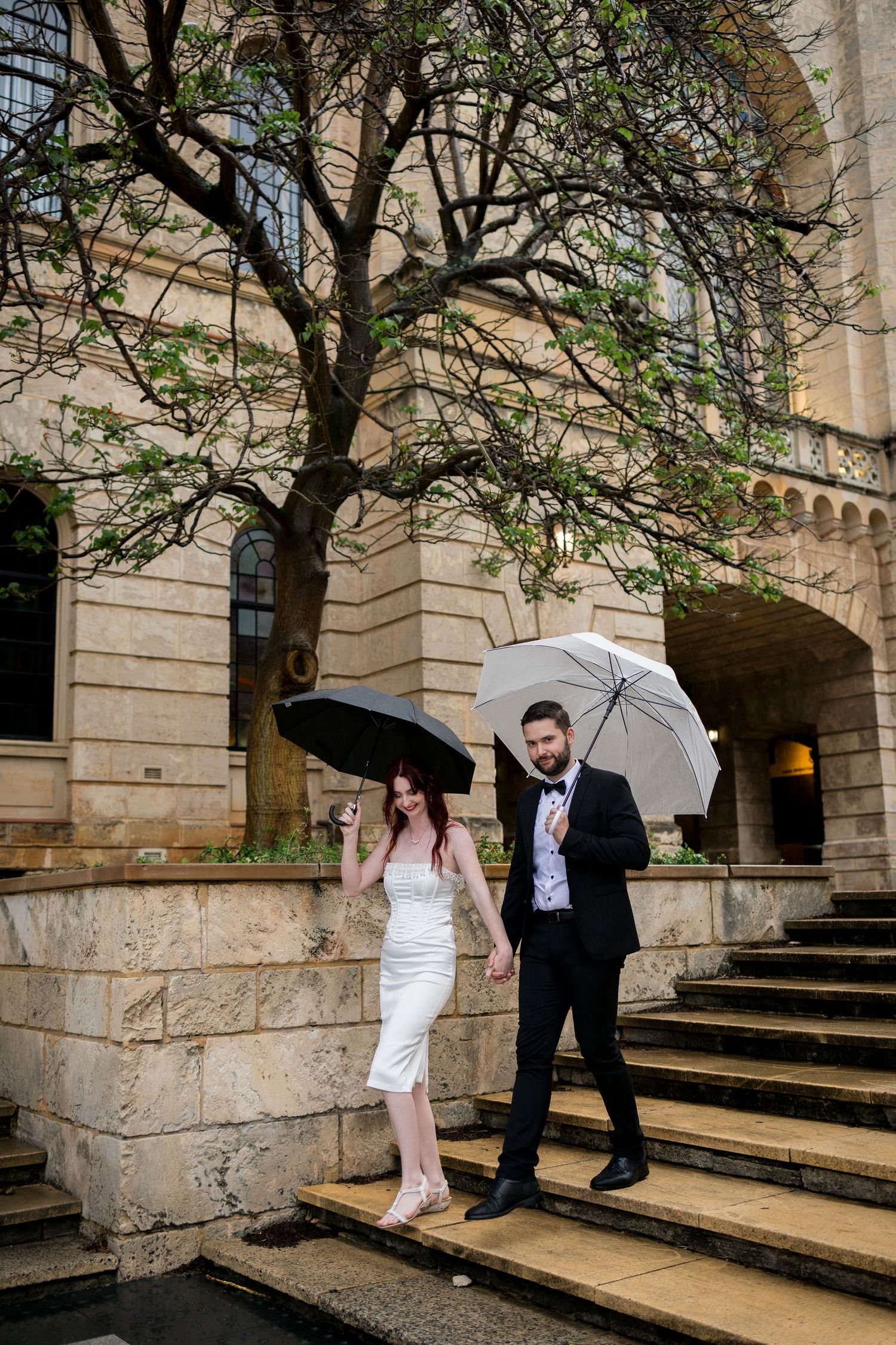 Couple descends stone steps with umbrellas at historic building entrance.