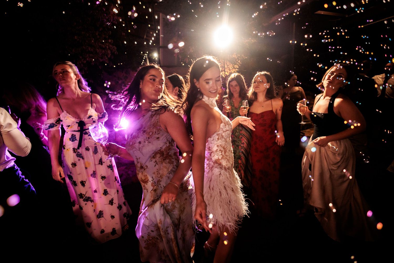 Guests in elegant gowns dance and celebrate at a nighttime wedding reception with sparkling fireworks.