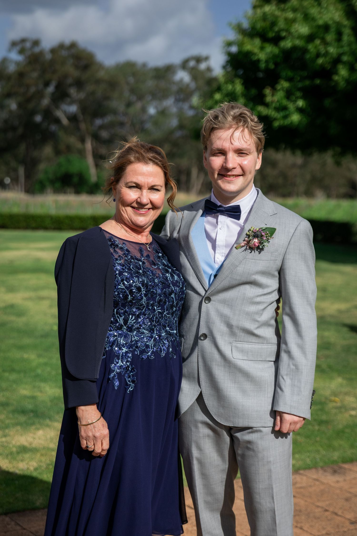 Young groom in grey suit with boutonniere poses with older woman in navy blue dress at outdoor wedding venue.