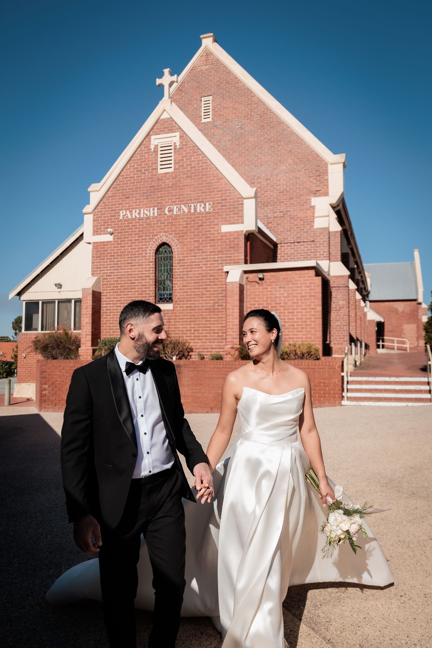 Bride and groom holding hands and smiling outside a red brick Parish Centre church on their wedding day.