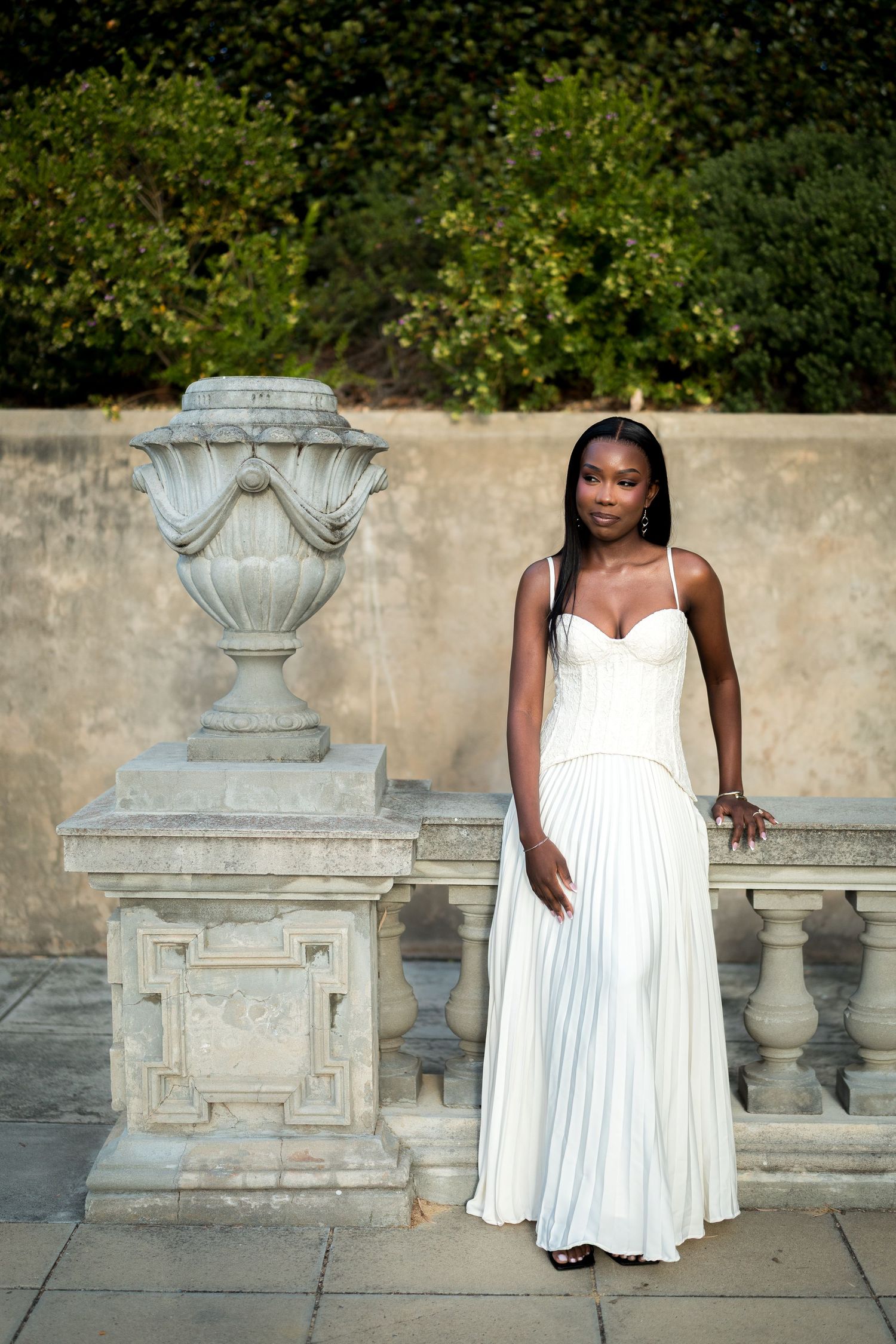 Woman in a flowing white pleated dress stands beside a carved stone urn and balustrade in a sunlit garden with green hedges.