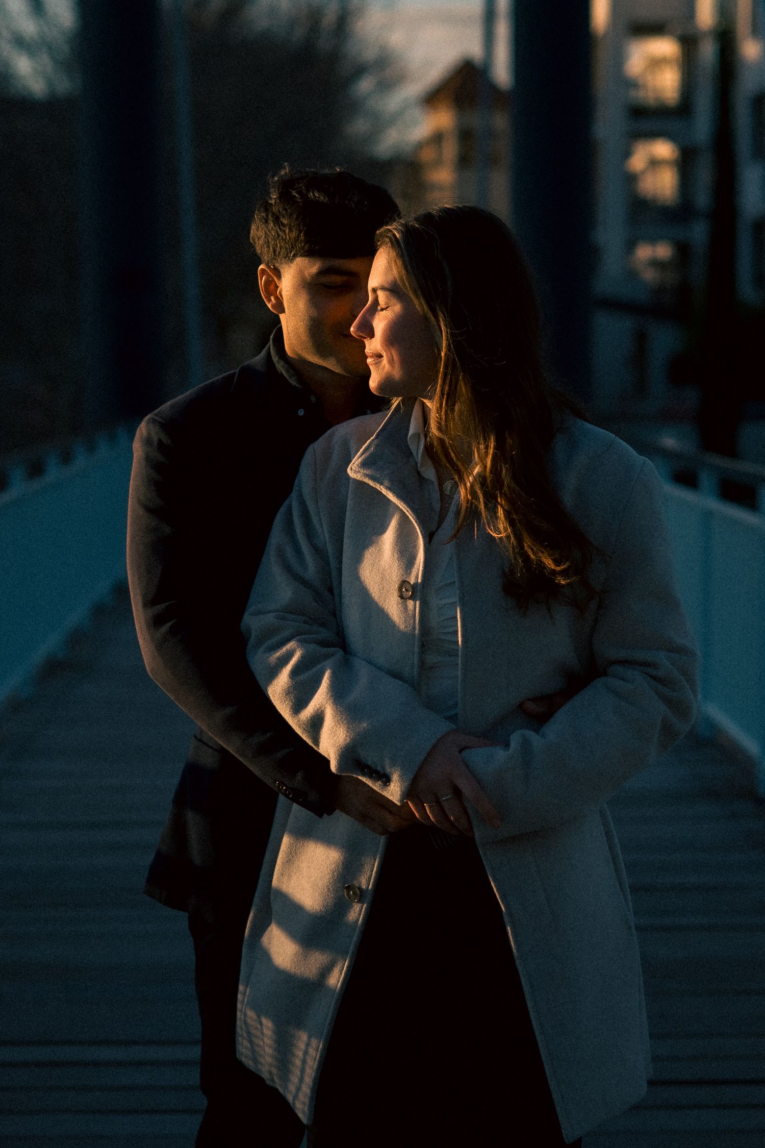 Couple embracing on a bridge at golden hour, woman in white coat smiling softly in warm evening light.