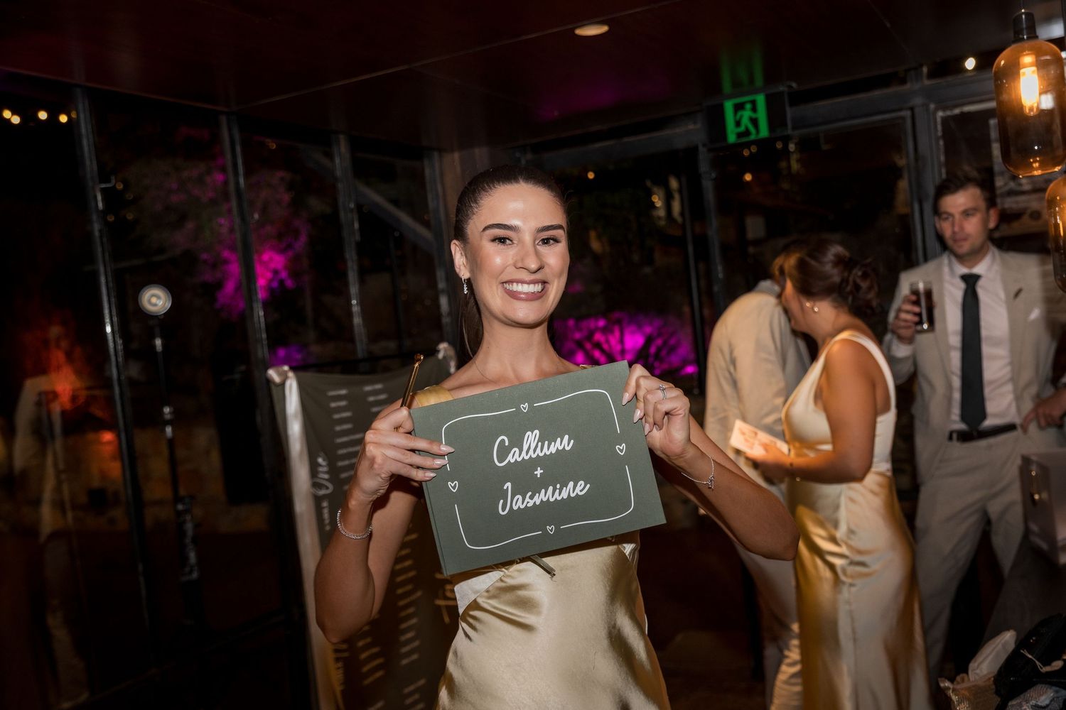 Woman in gold dress smiling and holding a chalkboard sign reading Callum and Jasmine at a wedding reception.