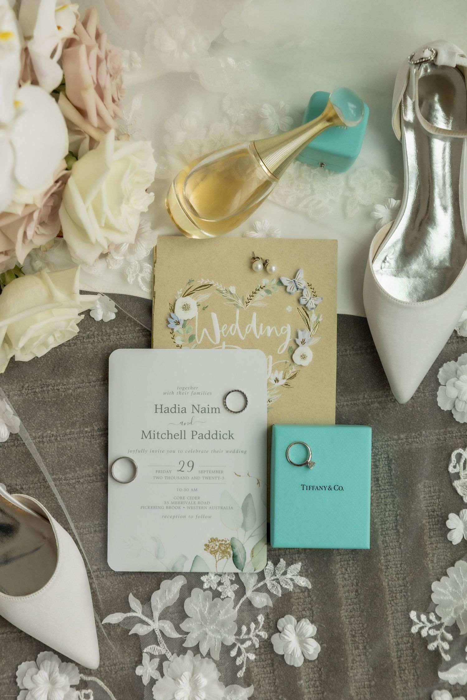 Elegant wedding flat lay featuring Tiffany & Co. ring box, floral invitations, white heels, gold perfume, and bridal bouquet.