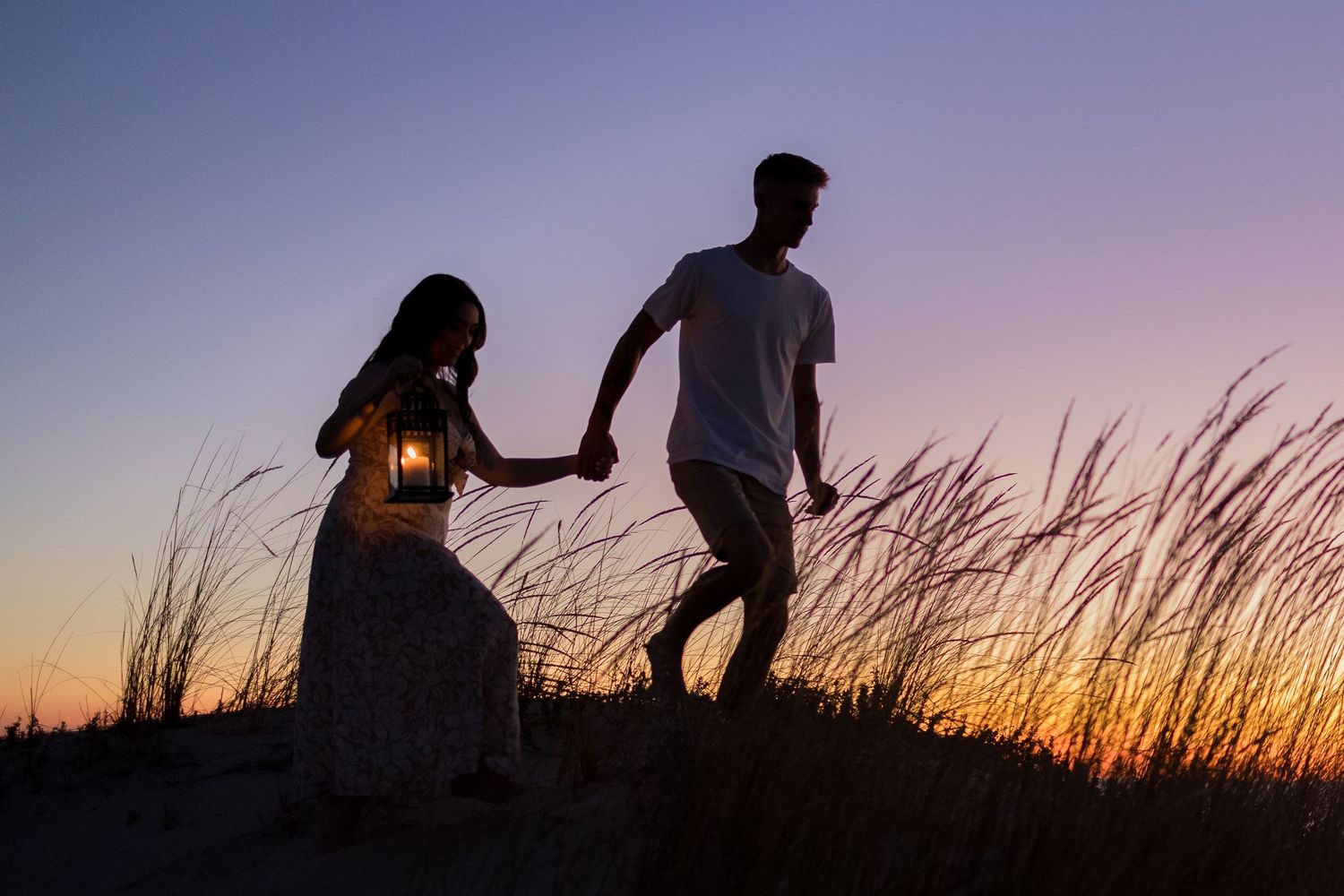 Silhouette of a couple holding hands at sunset in tall grass, woman carrying a glowing lantern.