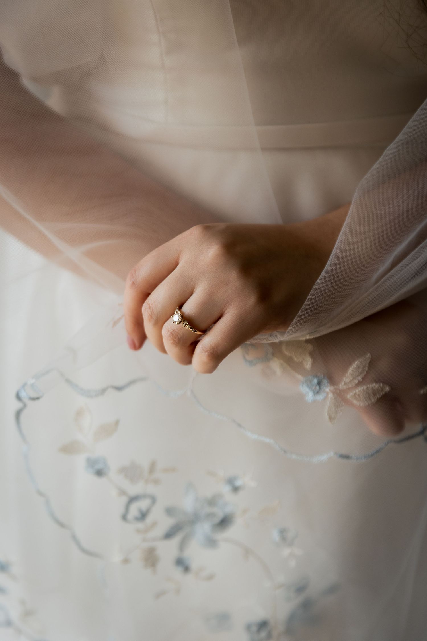 Close-up of bride's hand wearing gold diamond engagement ring on embroidered floral wedding dress.