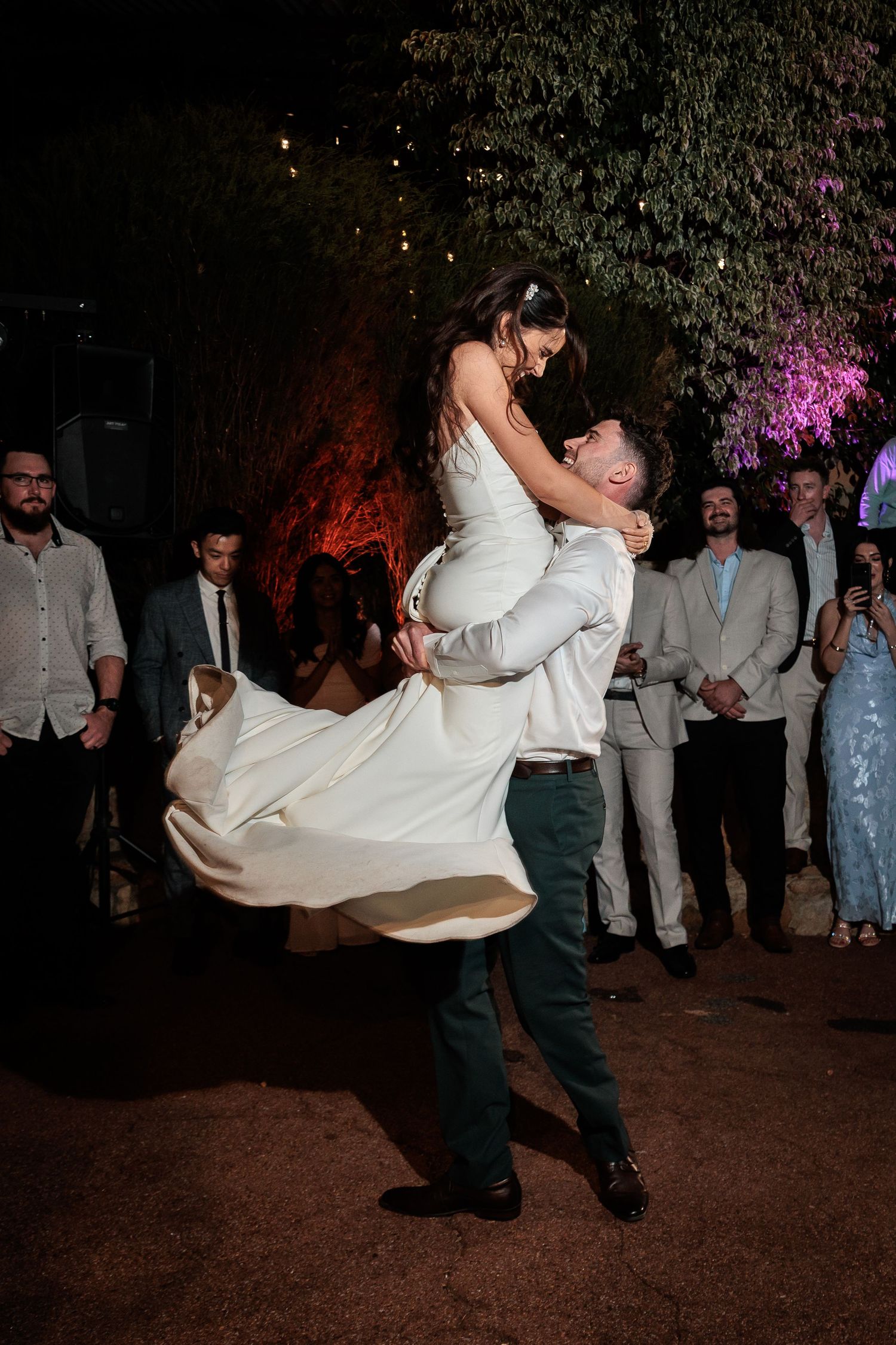 Groom lifts bride during first dance at outdoor wedding reception with guests watching at night.