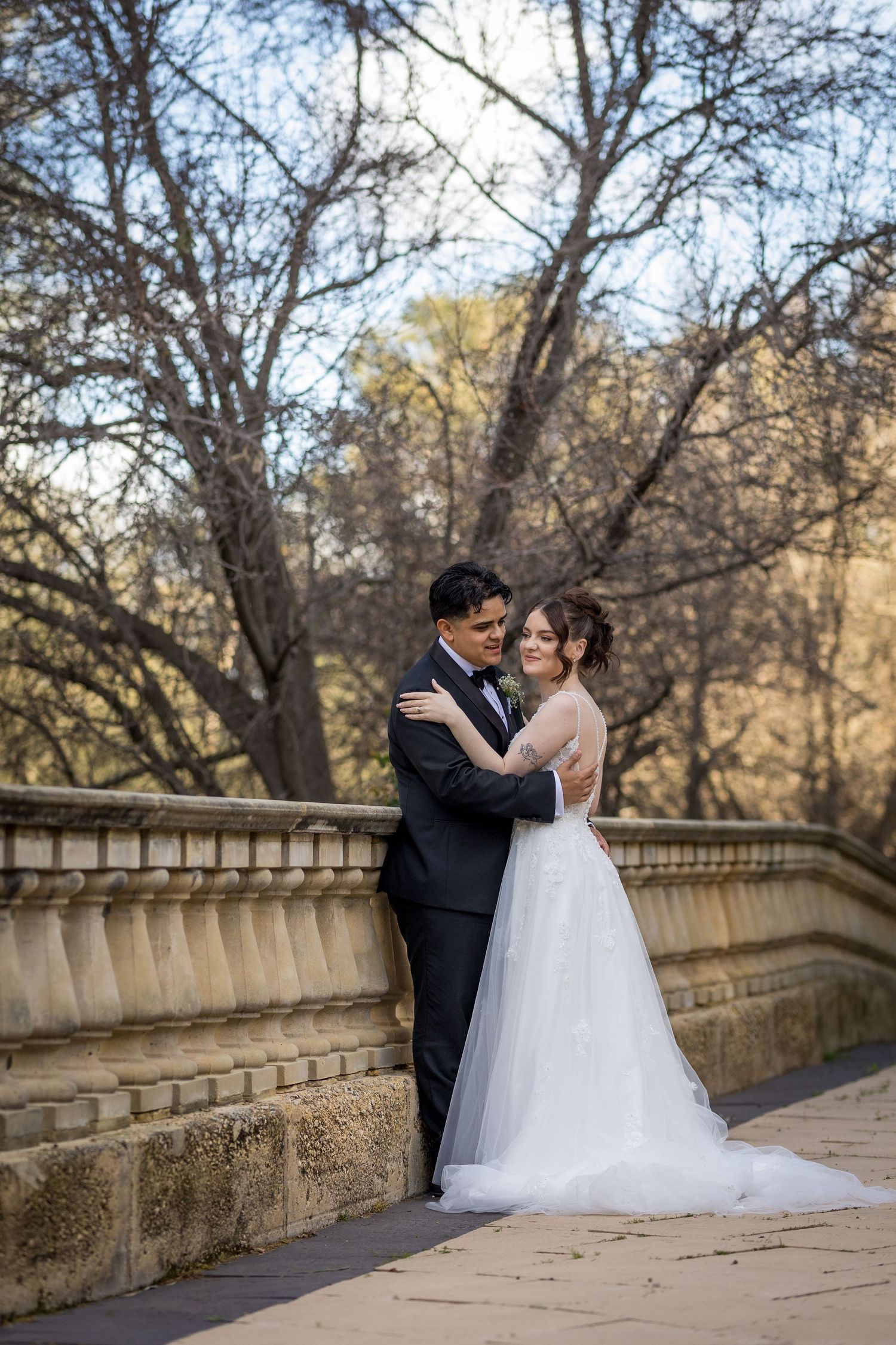 Bride and groom embrace on a stone bridge surrounded by bare trees in an outdoor wedding portrait.