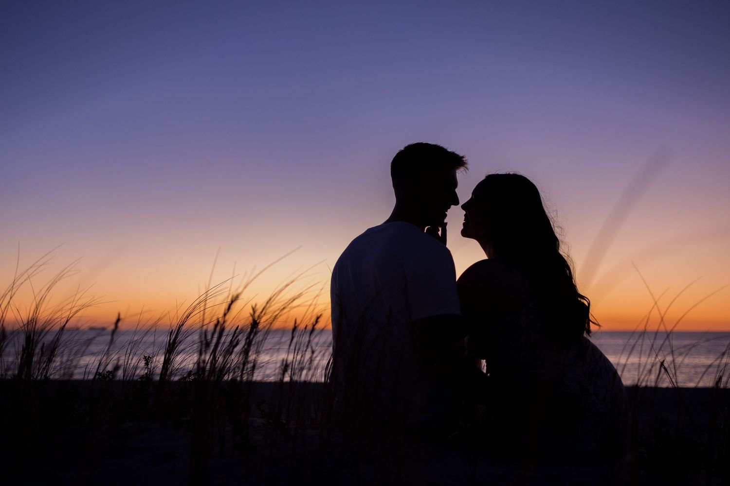 Romantic couple silhouette facing each other against a vibrant ocean sunset with tall grass in foreground.