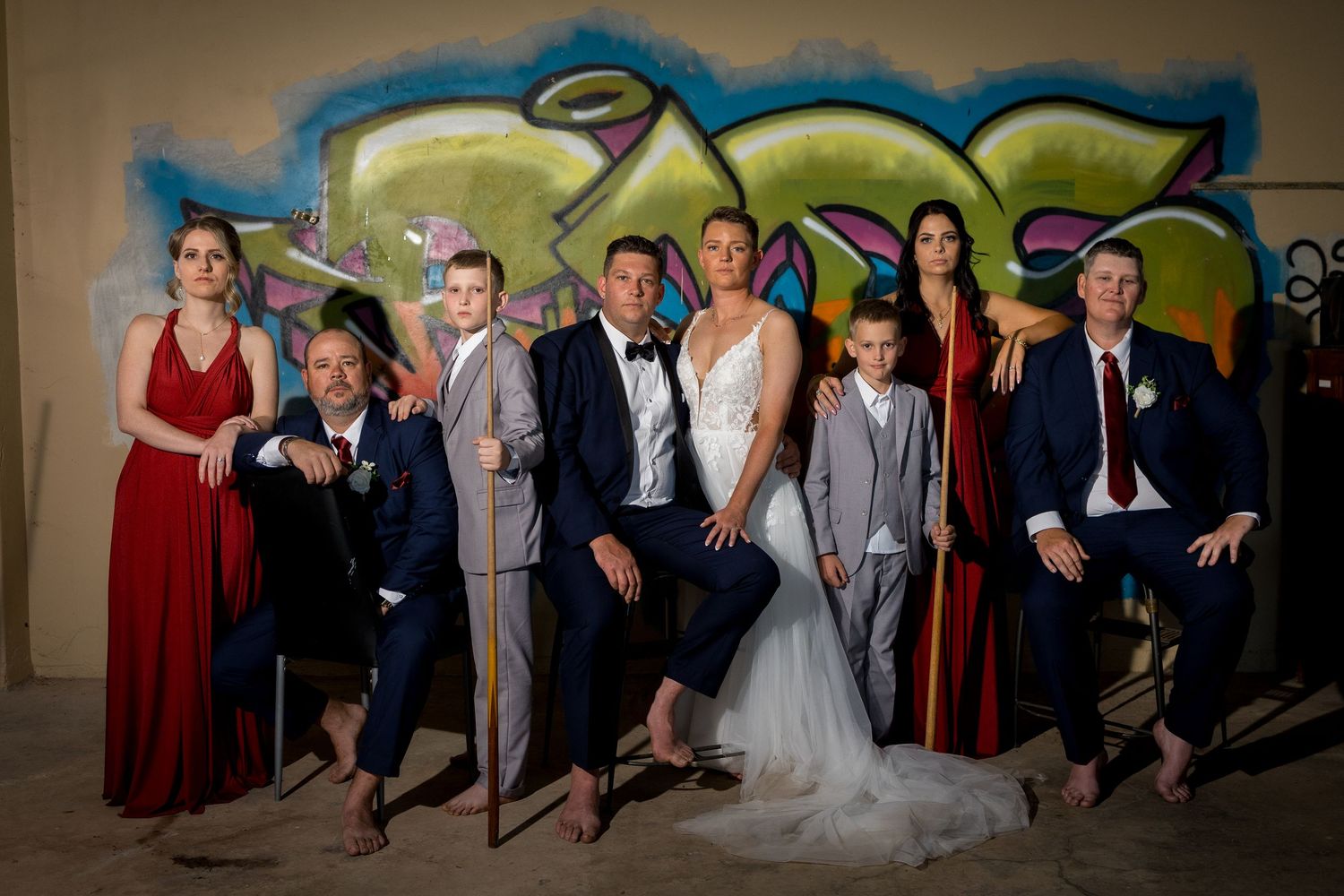 Wedding party poses playfully against blue wall in formal attire with bridesmaids in burgundy dresses.