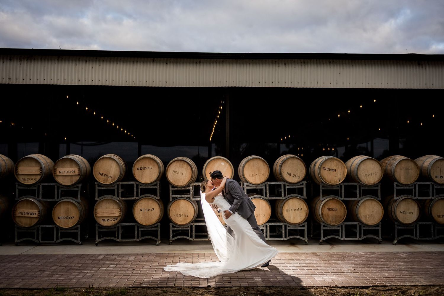 Bride and groom share a romantic dip kiss in front of rustic wine barrels at a winery wedding venue.