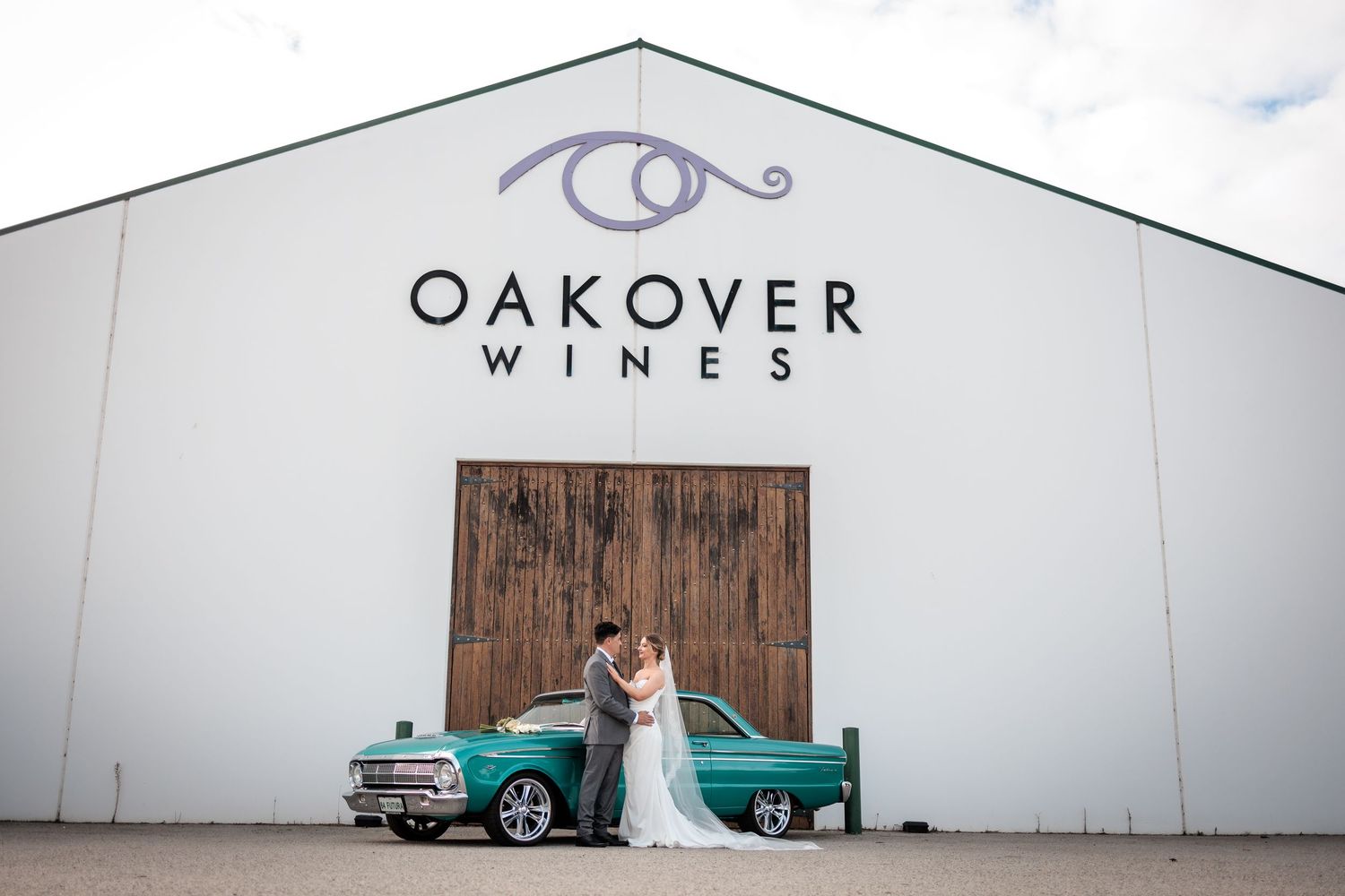 Bride and groom embrace in front of Oakover Wines white barn with a vintage teal car, in Swan Valley, Western Australia.