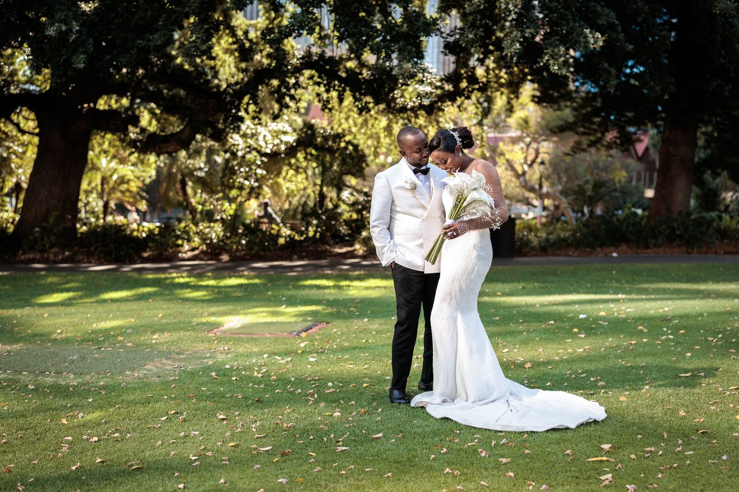 Bride and groom sharing a romantic moment on lush green lawn beneath large oak trees.