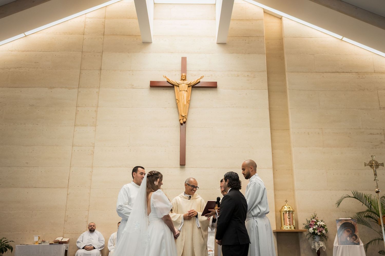 Couple exchanging vows at a Catholic church wedding ceremony with a priest and crucifix on the wall.