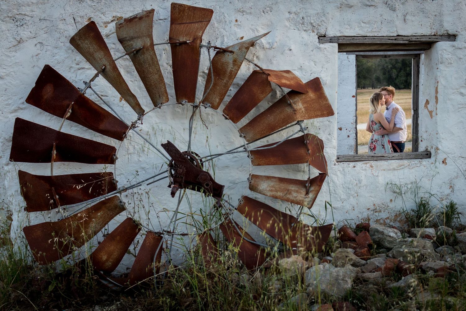 Couple embracing in window of rustic white building beside large vintage windmill wheel in outdoor engagement photo.