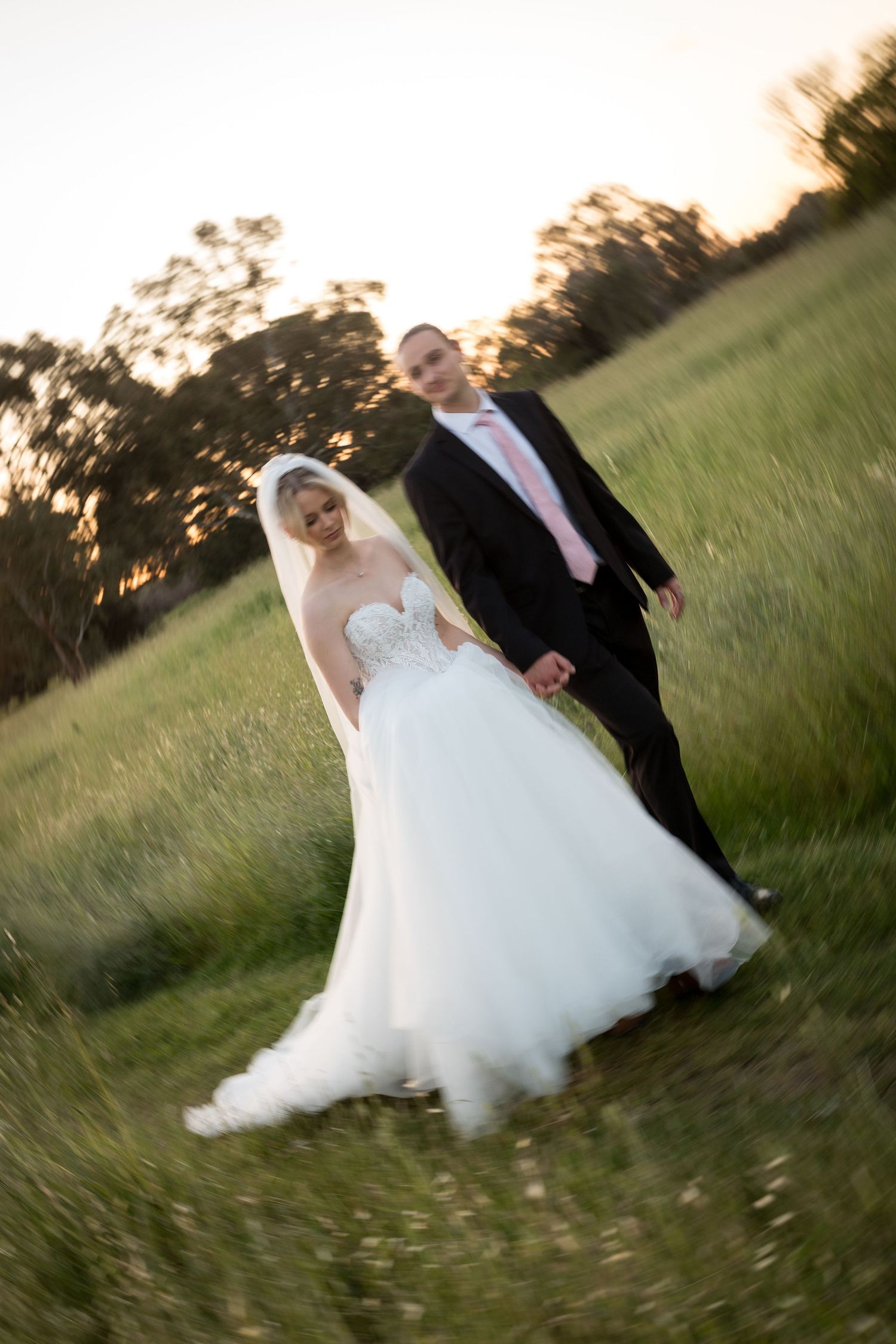 Bride and groom walk hand-in-hand through a lush green field at sunset, bride in white gown, groom in black suit.