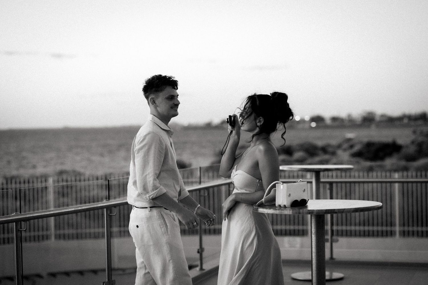 Black and white photo of a couple holding hands on a waterfront terrace at sunset, woman drinking from a glass.