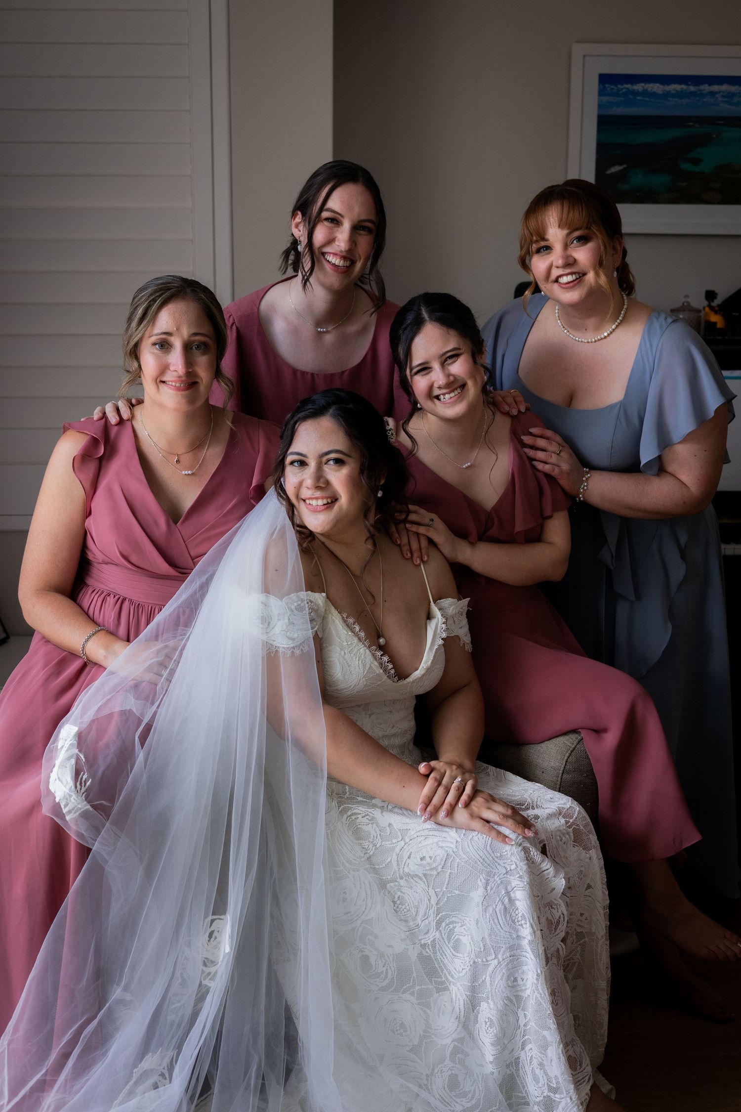 Bride in white lace gown and veil poses with four bridesmaids in pink and blue dresses before the wedding ceremony.