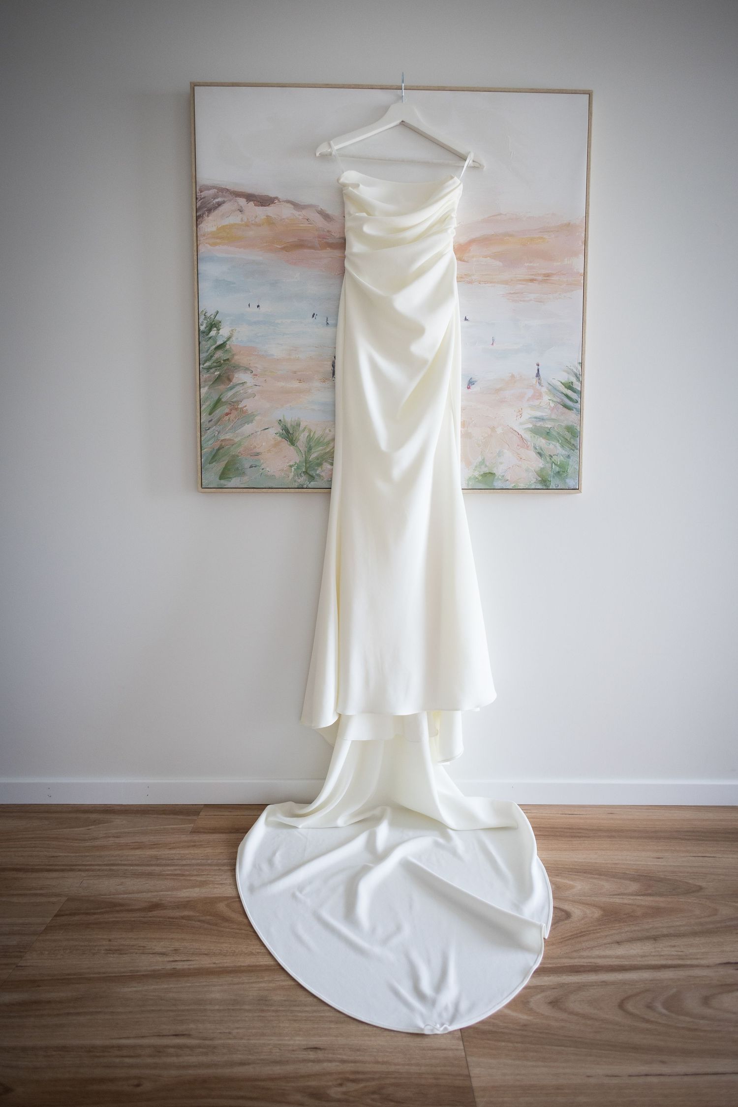 Elegant ivory satin wedding dress with a long train hanging on a white hanger against a pastel landscape painting.