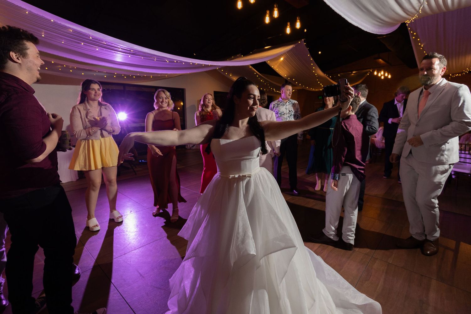 Wedding guests dancing and celebrating on a dance floor illuminated with purple lighting at an evening reception.