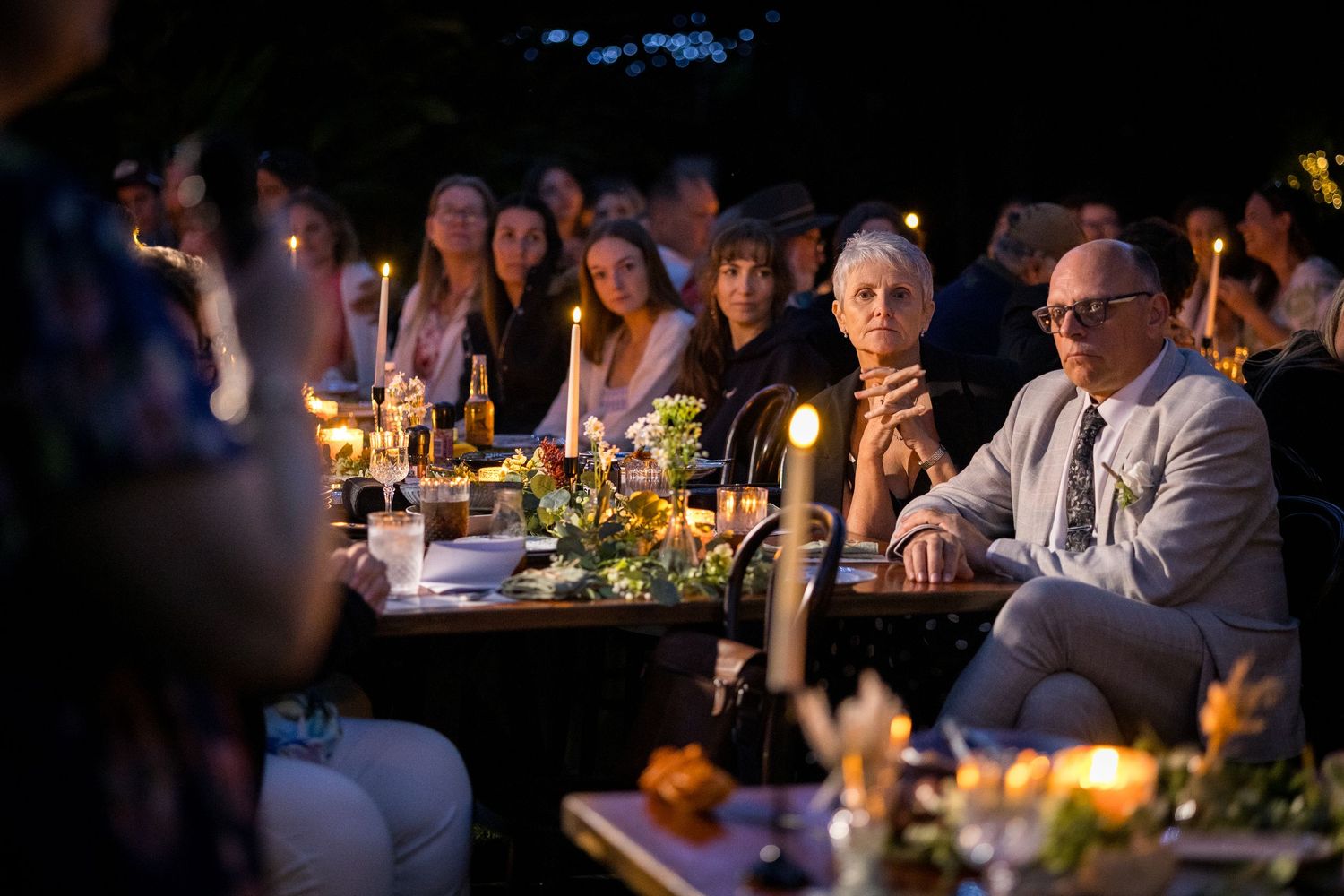 Wedding guests enjoy an intimate candlelit outdoor reception dinner under string lights at dusk.