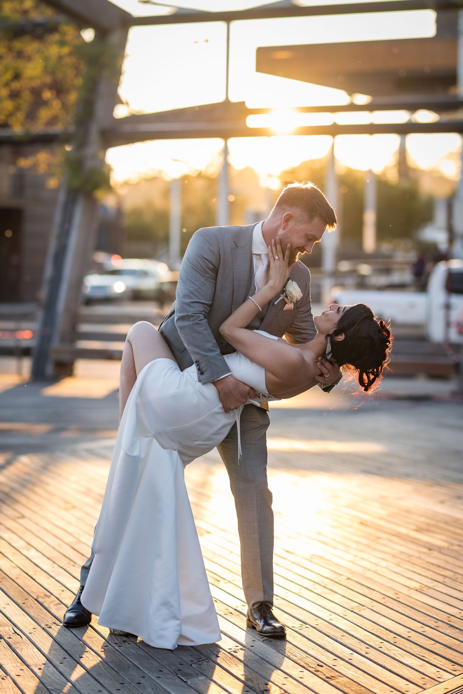 Groom dips bride in a romantic sunset kiss outdoors, both dressed in elegant wedding attire on a wooden deck.