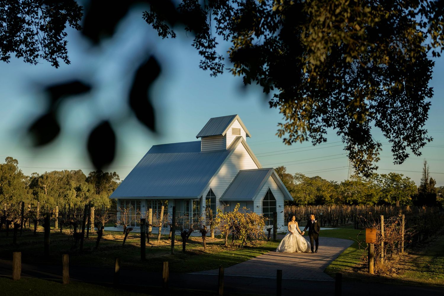 Bride and groom walk toward a white chapel at golden hour, framed by lush trees at a vineyard wedding venue.