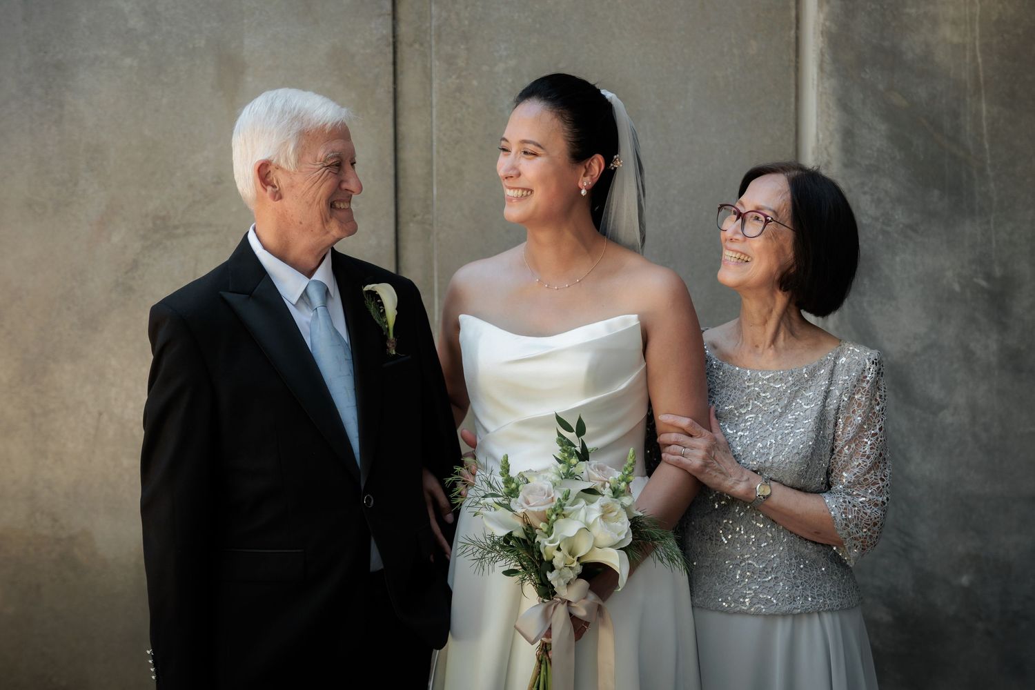 Bride in white gown holds bouquet flanked by proud parents in formal attire against gray wall.