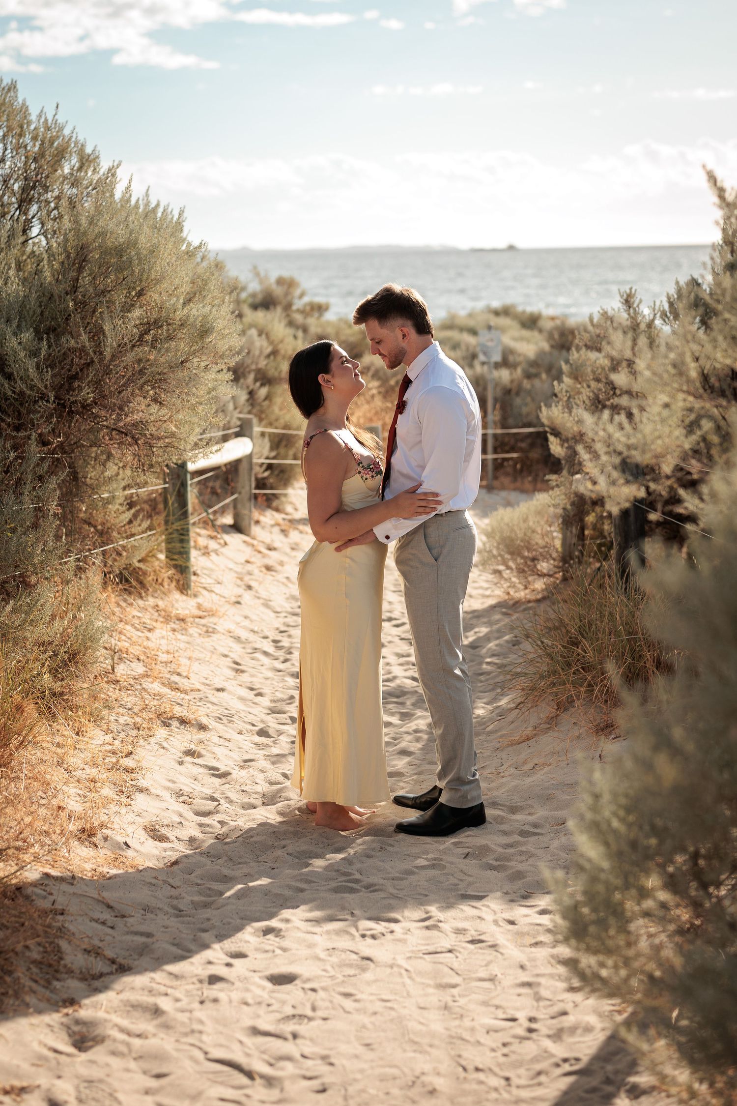 Couple sharing a romantic moment on a sandy beach path surrounded by coastal shrubs, woman in yellow dress, man in white shirt.