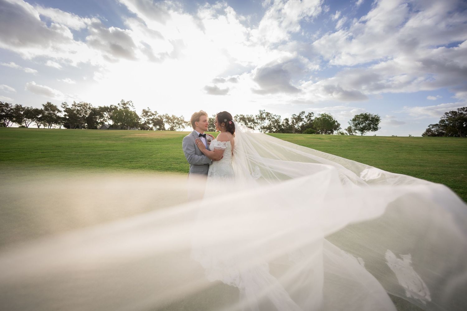 Bride and groom kiss on a green golf course as her dramatic cathedral veil flows in the wind under blue skies.