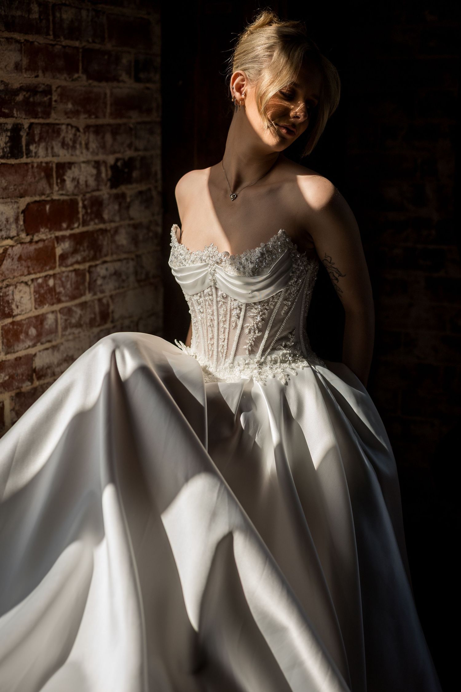 Bride in strapless corset ballgown poses in sunlight against rustic brick wall.