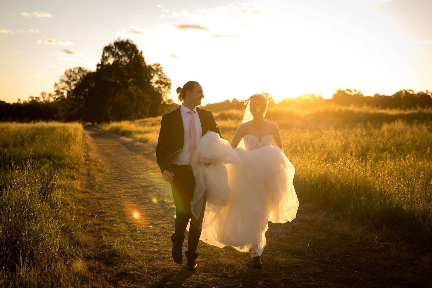 Bride and groom running through golden sunset meadow path in wedding attire, romantic outdoor ceremony photo.