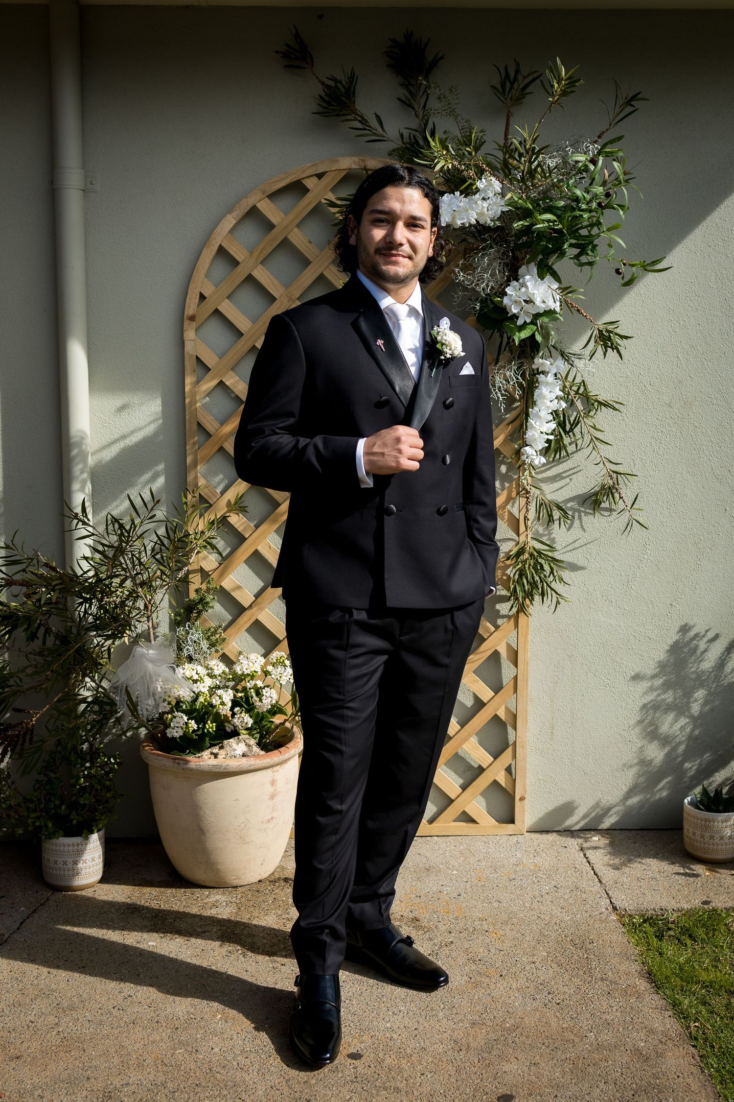 Groom in black double-breasted suit with white boutonniere poses before wooden trellis with greenery and flowers.