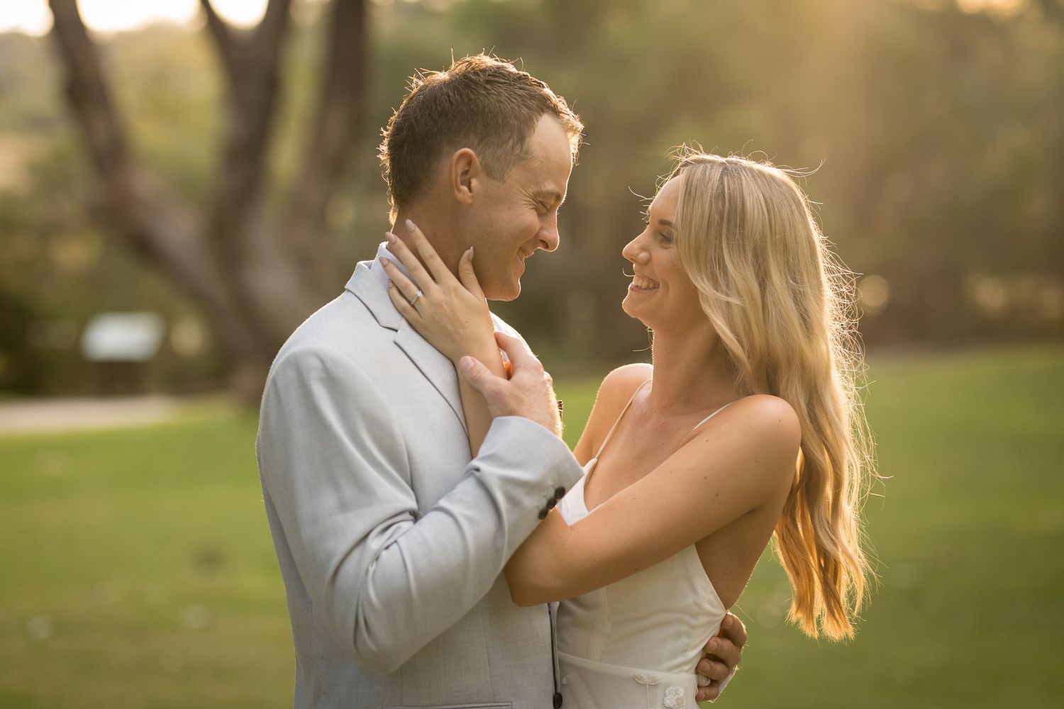 A smiling couple embraces outdoors at golden hour, with the woman in a white dress and man in a light gray suit.