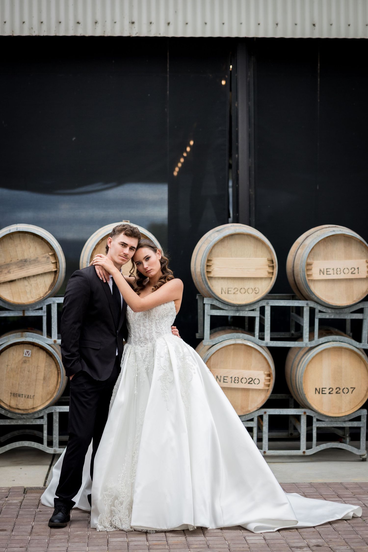 Bride and groom posing elegantly in front of stacked wooden wine barrels at a winery wedding venue.