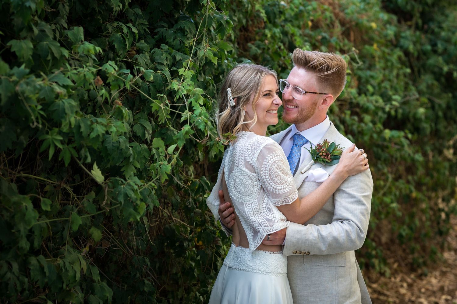 A bride and groom embrace and smile together outdoors in front of lush green foliage on their wedding day.