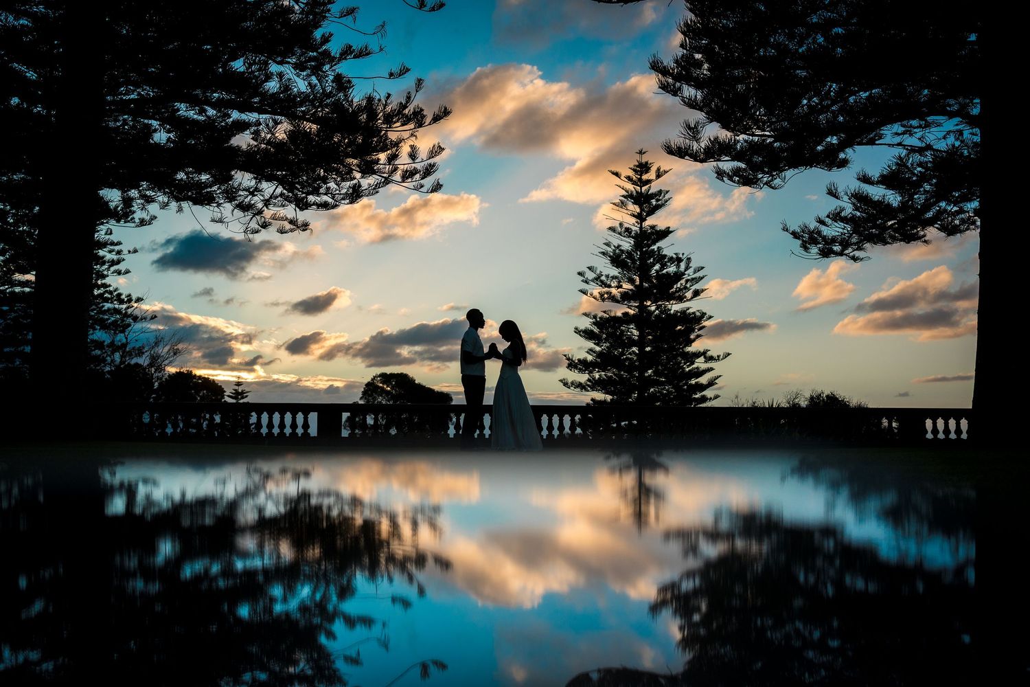 Silhouetted couple embracing on a terrace at sunset, with evergreen trees and a colorful cloudy sky reflected in a still pool below.
