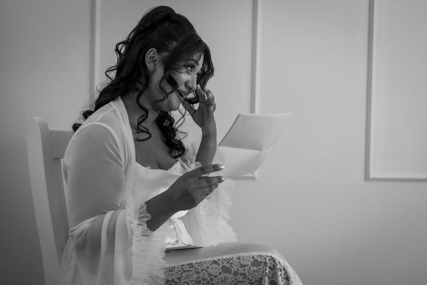Black and white photo of a bride in white dress sitting, examining herself in a small mirror before her wedding.