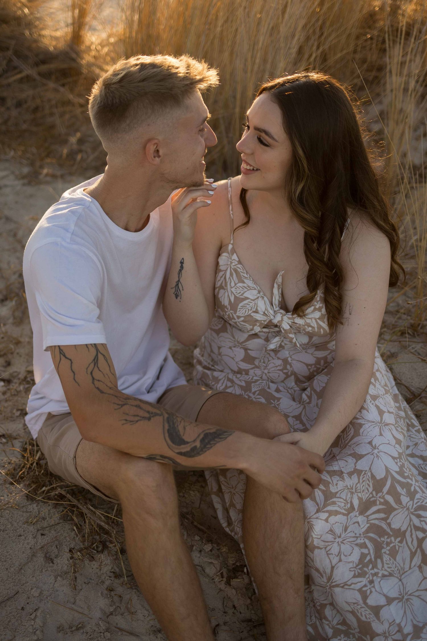 Tattooed couple sitting on beach dunes at sunset, smiling at each other in romantic moment.