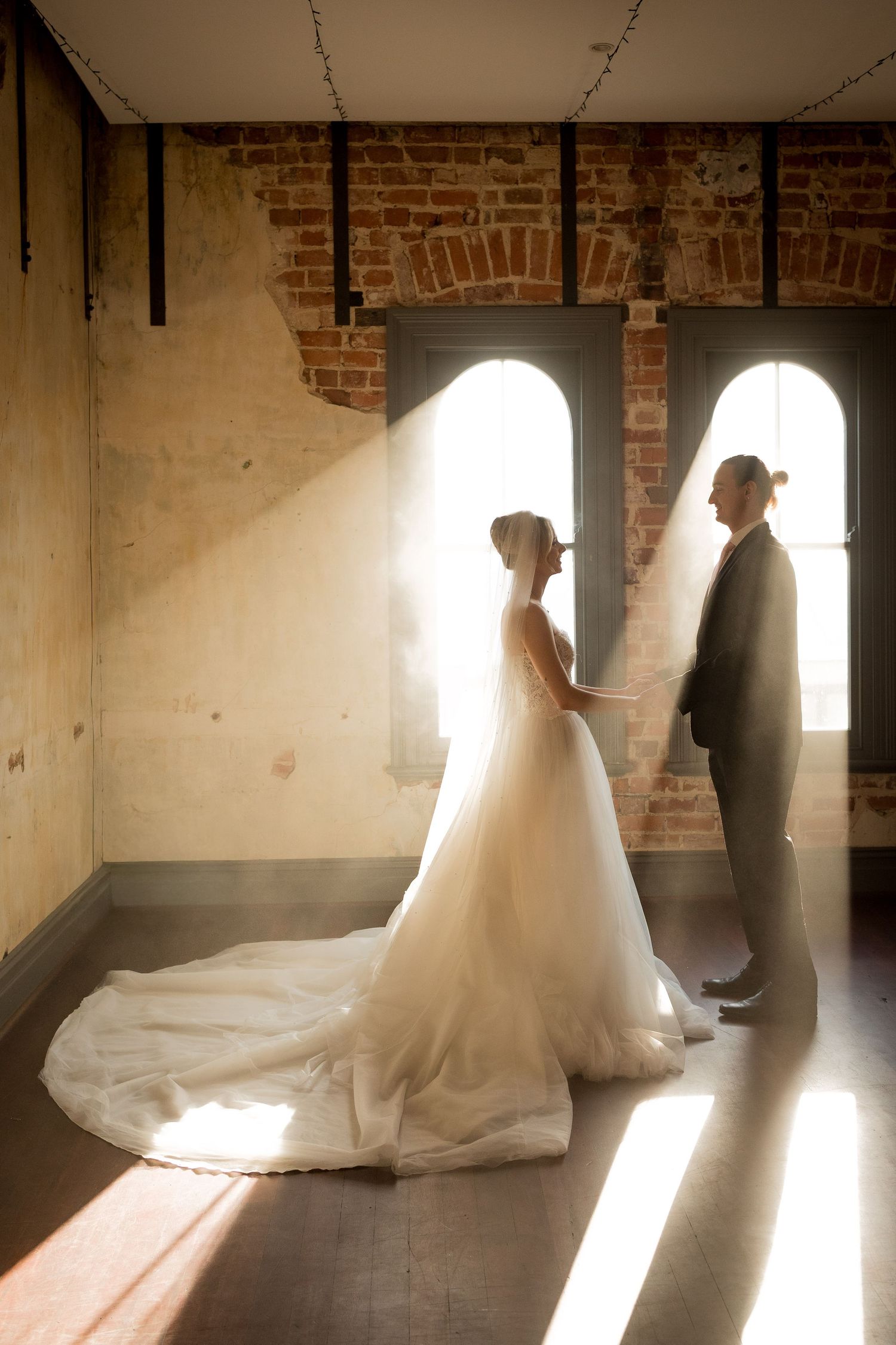 Bride and groom share a romantic moment in soft sunlight by arched brick windows in an elegant wedding venue.