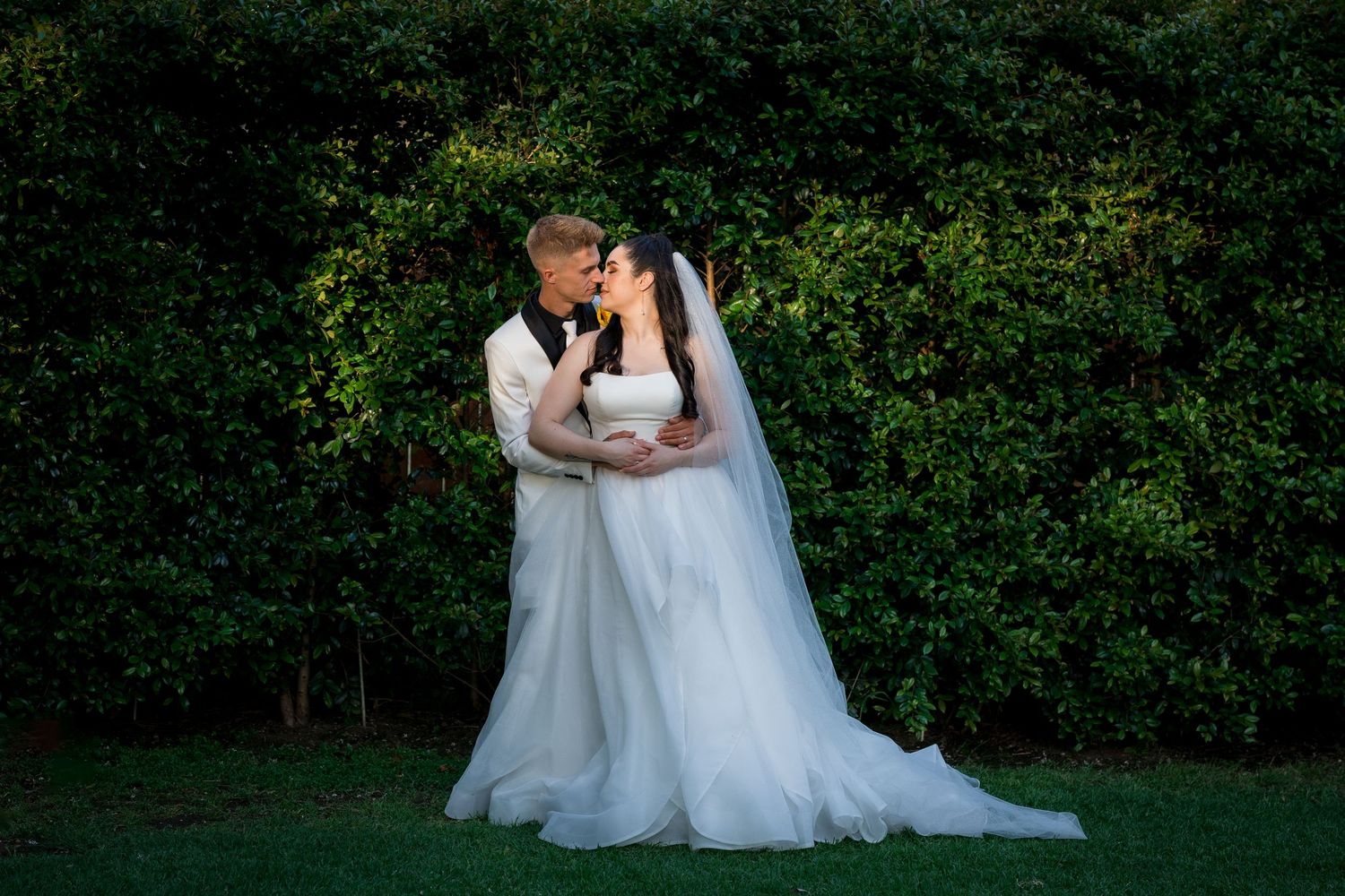 Bride and groom sharing a romantic kiss in front of lush green hedges during outdoor wedding ceremony.