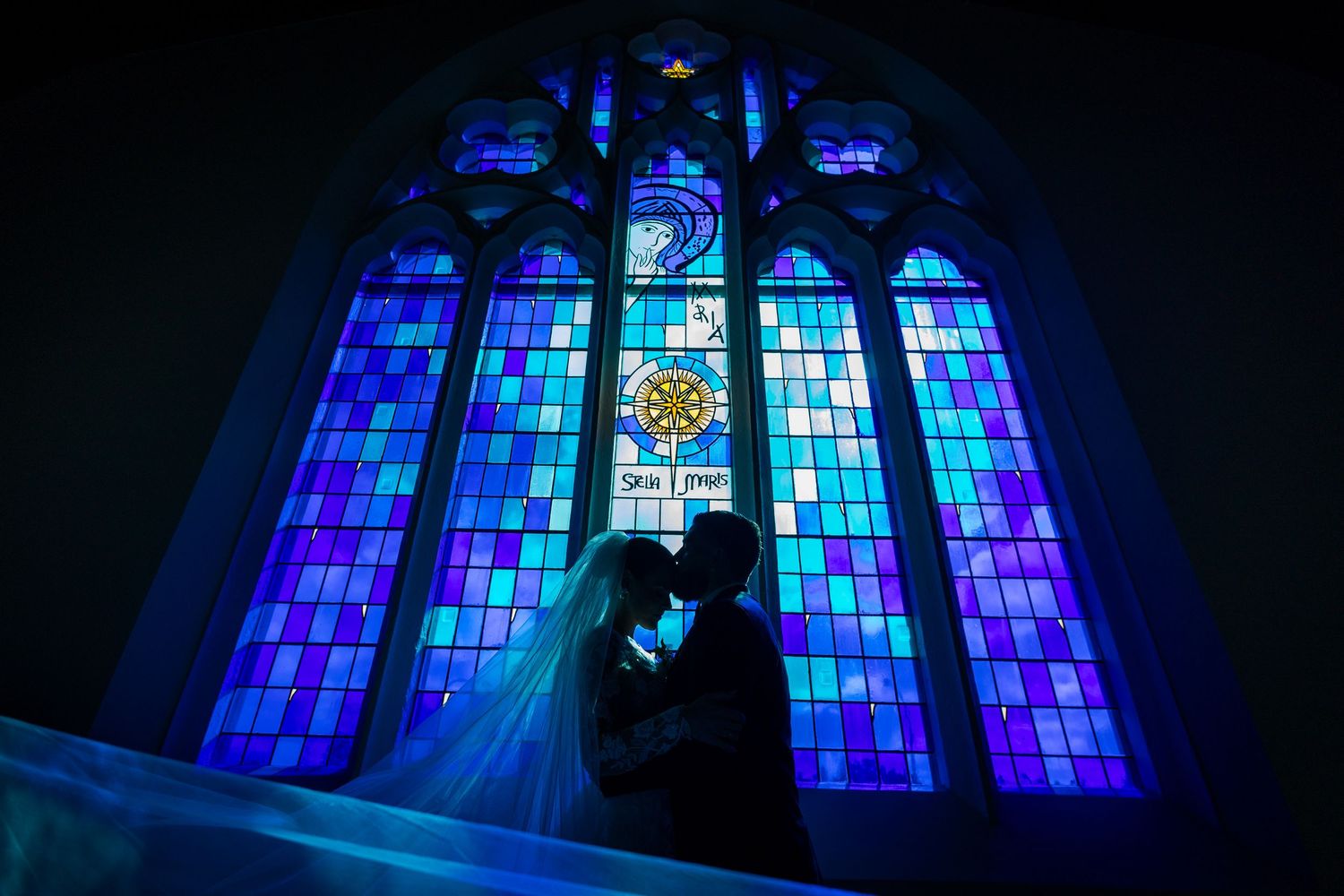 Bride and groom silhouetted against stunning blue stained glass church window.