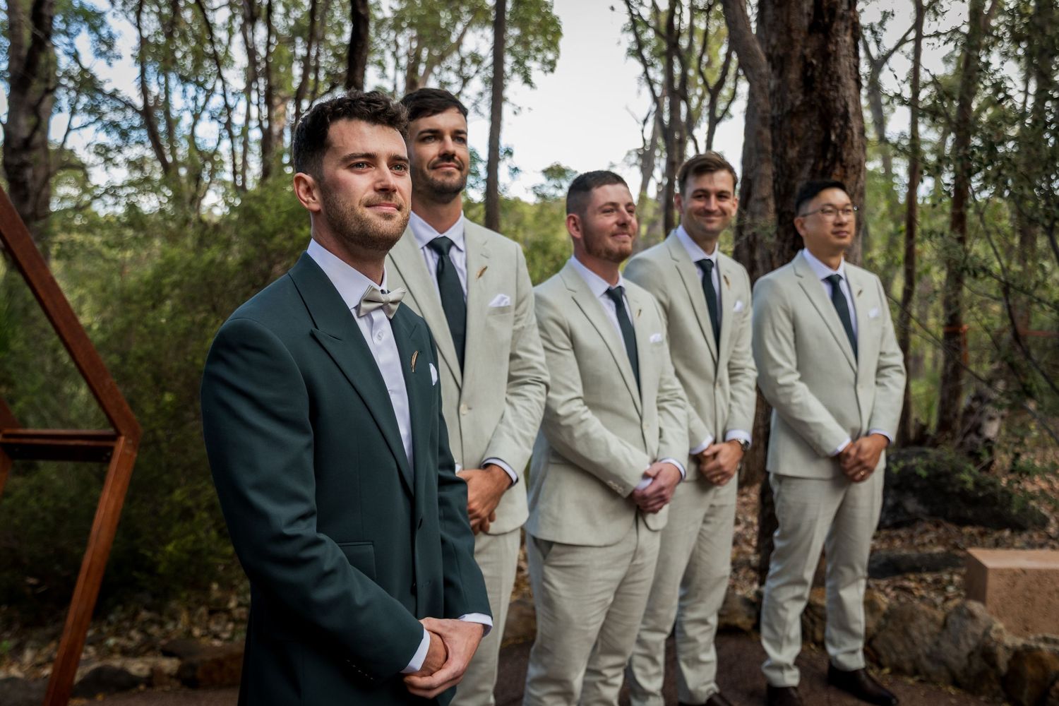 Groom in dark suit stands with groomsmen in tan suits at outdoor woodland wedding ceremony.