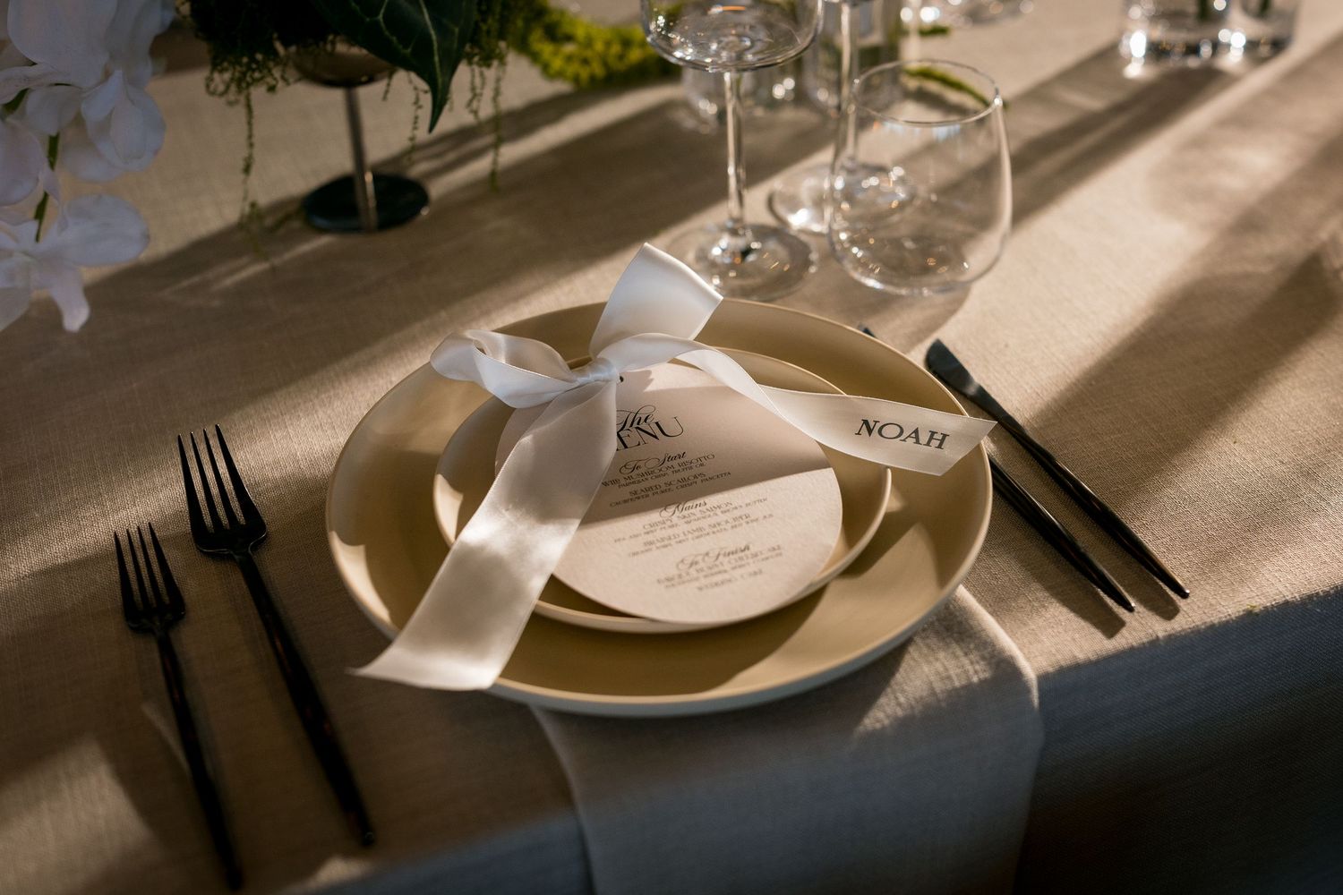 Elegant wedding place setting with gold plates, white ribbon bow, menu card, black cutlery, and crystal glasses on a neutral tablecloth.