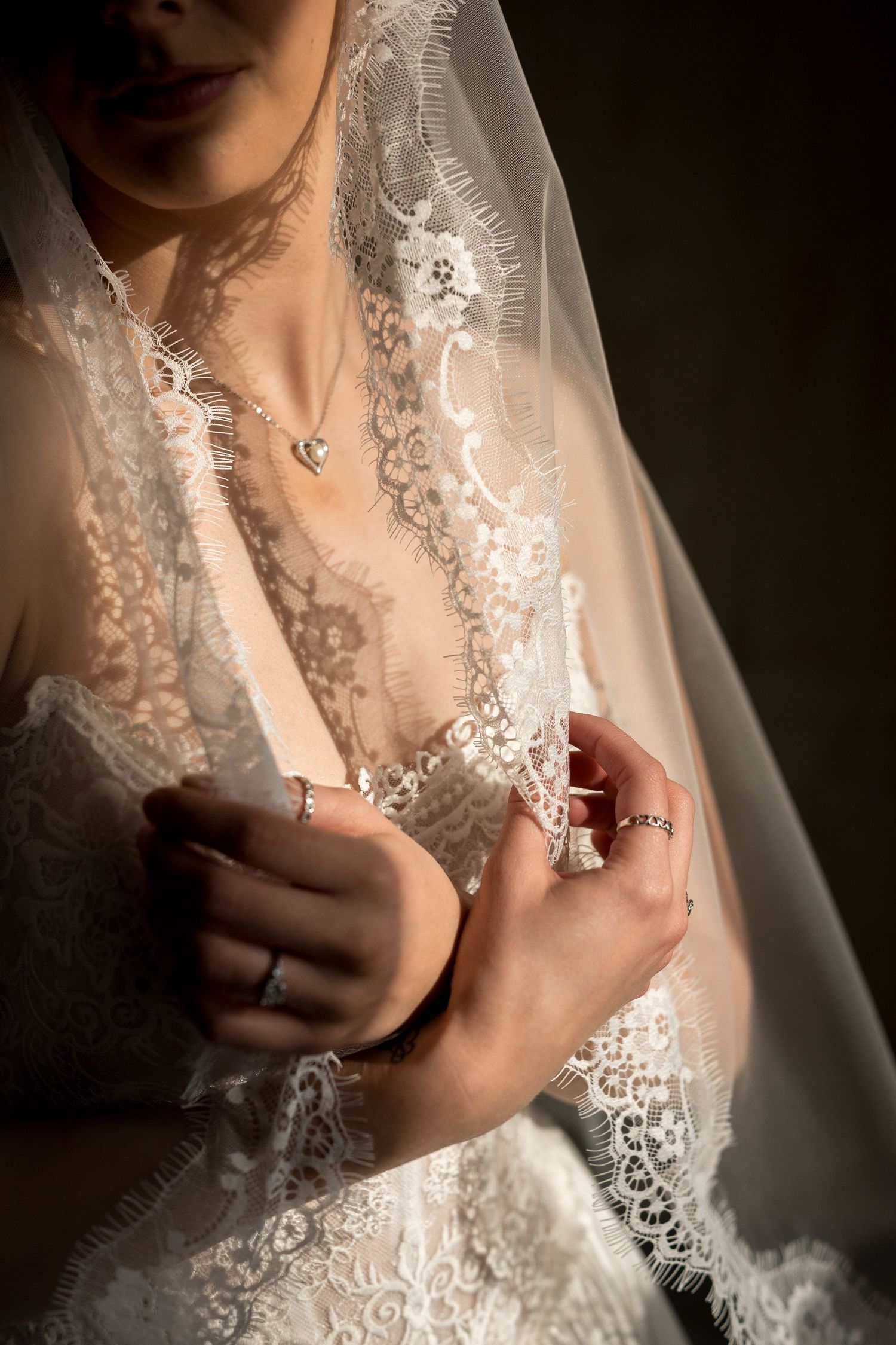 Bride in lace wedding dress and veil adjusting gown, wearing diamond ring and heart necklace.